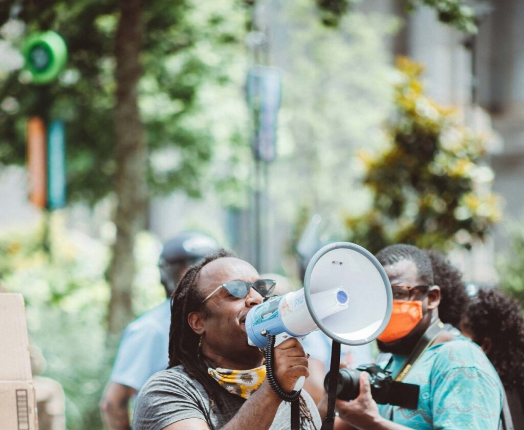 A Black man with a megaphone addresses a crowd at an outdoor voter organizing event