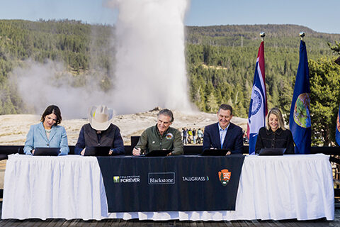 Five fund partners seated at the signing table for the Conservation and Stewardship Fund, with Old Faithful erupting behind them. Photo by the National Park Service.
